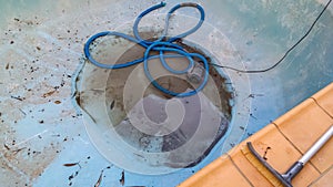 Bottom of drained swimming pool with just a little dirty water around the drain and a vacuum hose curled nearby with section of
