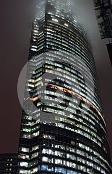 A bottom-up view of the facade of a modern office building at night in fog. Light of the night city. Moscow City