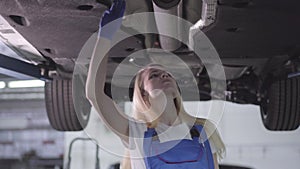 Bottom view of confident caucasian auto mechanic checking fixed automobile. Blond young woman working in car repair shop