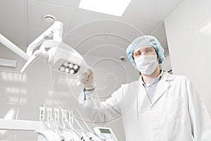 Bottom view of a doctor portrait dentist, surgeon in uniform and mask on his face, wearing glasses, holding lamp in her