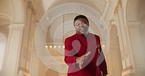 Bottom view of gospel singer in red suit looking to camera and singing while standing at church hall. Young guy clapping