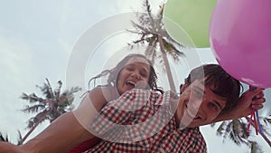 Bottom view of happy girl on the shoulders of her man holds balloons having fun on palm trees on the background. A