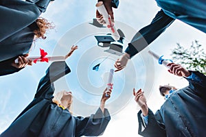 bottom view of happy multicultural graduates with diplomas throwing caps up with blue sky