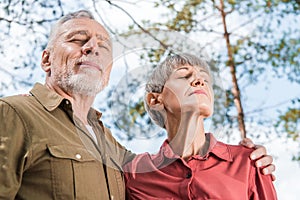 bottom view of happy senior couple embracing with closed eyes in forest.