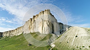 Bottom view of the high white rock on blue cloudy sky background. Shot. Sunny day and beautiful green meadow at the foot