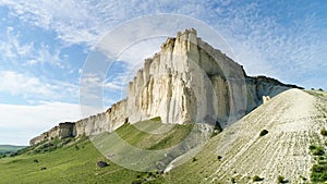 Bottom view of the high white rock on blue cloudy sky background. Shot. Sunny day and beautiful green meadow at the foot