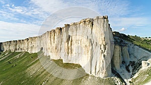 Bottom view of the high white rock on blue cloudy sky background. Shot. Sunny day and beautiful green meadow at the foot