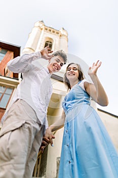 Bottom view of lovely young attractive couple having fun outdoors in the city, waving and smiling to camera and holding