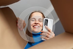 Bottom view of smiling happy Caucasian woman receiving online order, unpacking her carton parcel at home, being satisfied with