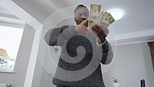 Bottom view of wealthy rich African American satisfied man scattering money standing in living room at home. Smiling