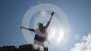 Bottom of view on young man standing on the edge of rock rises his hands on the sky and the sun on background. slow