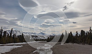 Boulder and gravel riverbed and snowcapped mountains under lenticular clouds in the spring in Denali National Park in Alaska