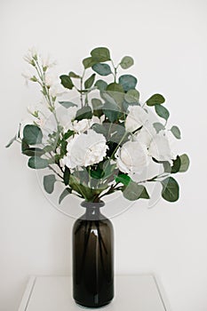 Bouquet of artificial plants and white flowers in vase, stands on light background.