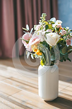 The bouquet of assorted flowers including white roses, pink ranunculus, and yellow blossoms, in a white ribbed vase, placed on a