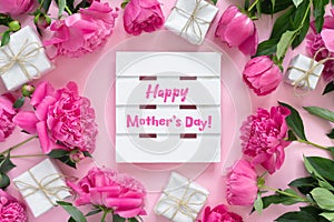 Bouquet of beautiful pink peonies with gift boxes in paper wrapping. Round frame of flowers. White wooden board with text Happy