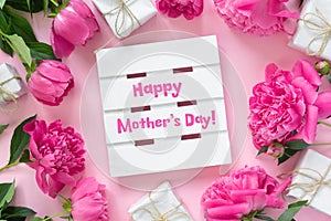 Bouquet of beautiful pink peonies with gift boxes in paper wrapping. Round frame of flowers. White wooden board with text Happy
