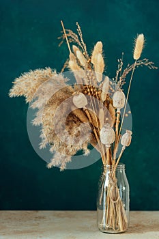 Bouquet of beige dried flowers in a glass vase on green-blue background.