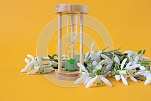 A bouquet of blooming snowdrops with an hourglass on a yellow background, a side view, a place for text-the concept of the arrival