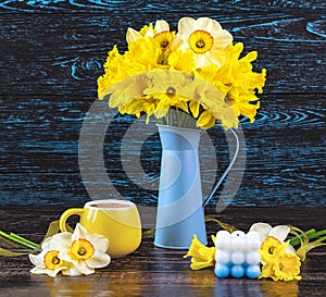 A bouquet of daffodil flowers in a blue vase, a yellow cup of coffee, a blue candle on a dark wooden backgroundGenerated image