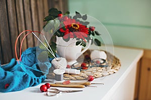 A bouquet of dogrose and red flower in a vase,  skeins, needles, scissors, knitwear,glasses, on a white table in the cozy home.