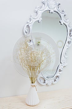 A bouquet of dry wildflowers stands in a white vase on a boudoir table by a vintage mirror