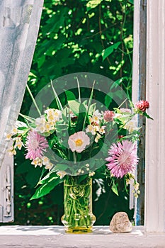 Bouquet of flowers of clover, cornflowers and Jasmine in a glass vase and a heart shaped stone on the windowsill of an open window