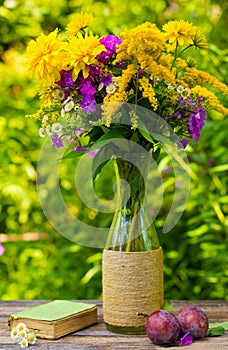 A bouquet of flowers of goldenrod, phlox chrysanthemums in a glass vase, ripe plums and old book