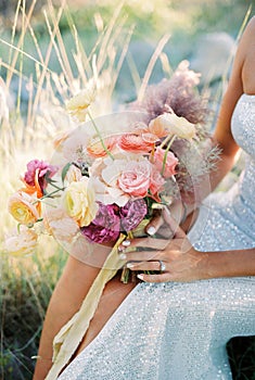 Bouquet of flowers in the hands of a bride sitting on a sunny lawn. Cropped. Faceless