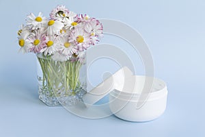 A bouquet of fresh flowers, daisies on green stems are in a glass with clean water and a white jar of cream on a blue background