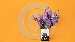 Bouquet of fresh lupine flowers in white vase shaped like bag and hello summer lettering with smile on black heart on