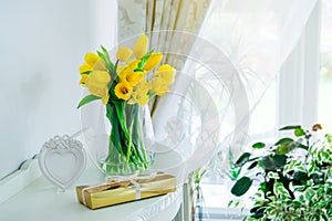 Bouquet of fresh yellow spring tulips and daffodils flowers, golden gift box and heart shaped frame on the chest drawers in light