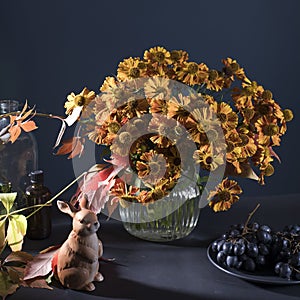 A bouquet of helenium with wild grape leaves in a fluted glass vase against a dark blue wall. Porcelain figurine of a hare