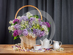 bouquet of lilacs in a basket on the table. cup of tea, teapot and milk jug. summer tea concept