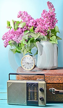Bouquet of lilacs in enameled kettle on antique suitcase, vintage radio, alarm clock on blue background. Retro style still life.
