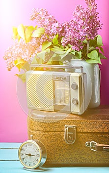 Bouquet of lilacs in enameled kettle on antique suitcase, vintage radio, alarm clock on pink background. Retro style still life.