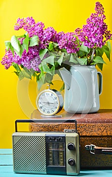 Bouquet of lilacs in enameled kettle on antique suitcase, vintage radio, alarm clock on yellow background. Retro style still life.