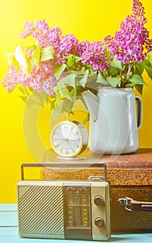 Bouquet of lilacs in enameled kettle on antique suitcase, vintage radio, alarm clock on yellow background. Retro style still life.