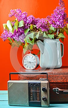 Bouquet of lilacs in enameled kettle on antique suitcase, vintage radio, alarm clock on yellow background. Retro style still life.