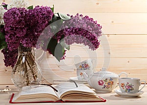 Bouquet of lilacs, open book, spectacles, teapot and cup of tea