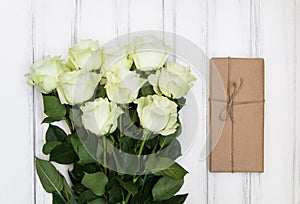 Bouquet of luxurious white roses and a present box in eco paper on white wooden background. Top view, flat lay