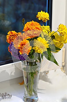 Bouquet of orange marigolds, blue aster and yellow flowers in a glass vase. The bouquet stands on a white window sill