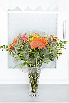 Bouquet of orange, yellow and red flowers in a vase in front of a window.