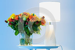 A bouquet of orange yellow and red roses flowers on a table next to a lamp with blue background close up view