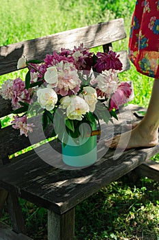 Bouquet of peony flowers in milk can, woamn standing on wooden garden bench