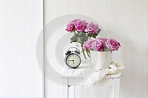 Bouquet of pink roses in a tin white bucket on an old stool with an alarm clock and a napkin with embroidery near the bed on a