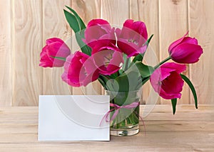 Bouquet pink tulips flowers in glass white vase with white paper card note with space for text on table on a wooden background