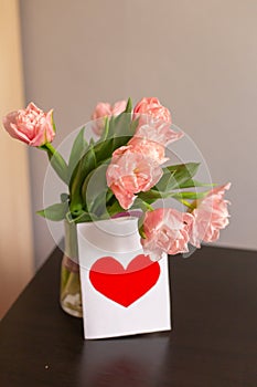 Bouquet of pink tulips in a glass vase with a postcard with a red heart. Romantic spring background with space for text.