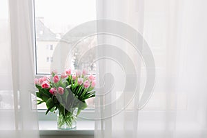 Bouquet of pink tulips in transparent vase on windowsill, against background of window. Flowers in interior.