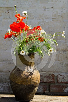 Bouquet of poppies and daisies in clay jug on wooden table against old brick wall