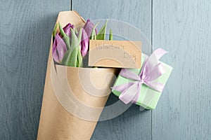 Bouquet of purple tulips in craft paper with gift on blue wood table with paper card for valentines day